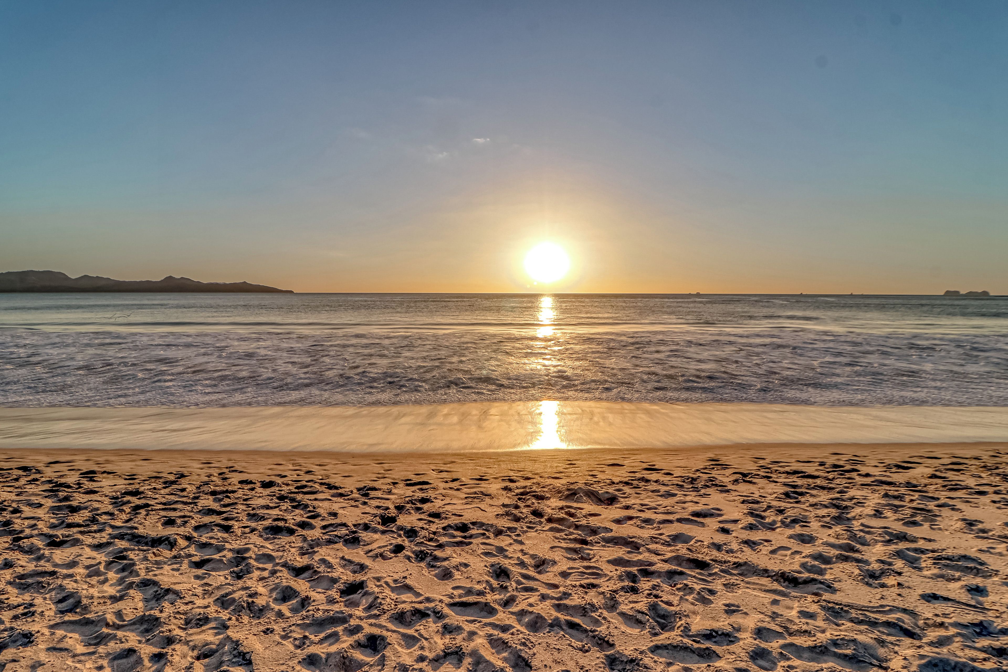 Una playa con huellas que conducen al agua, cielo despejado y el sol reflejándose en la superficie.