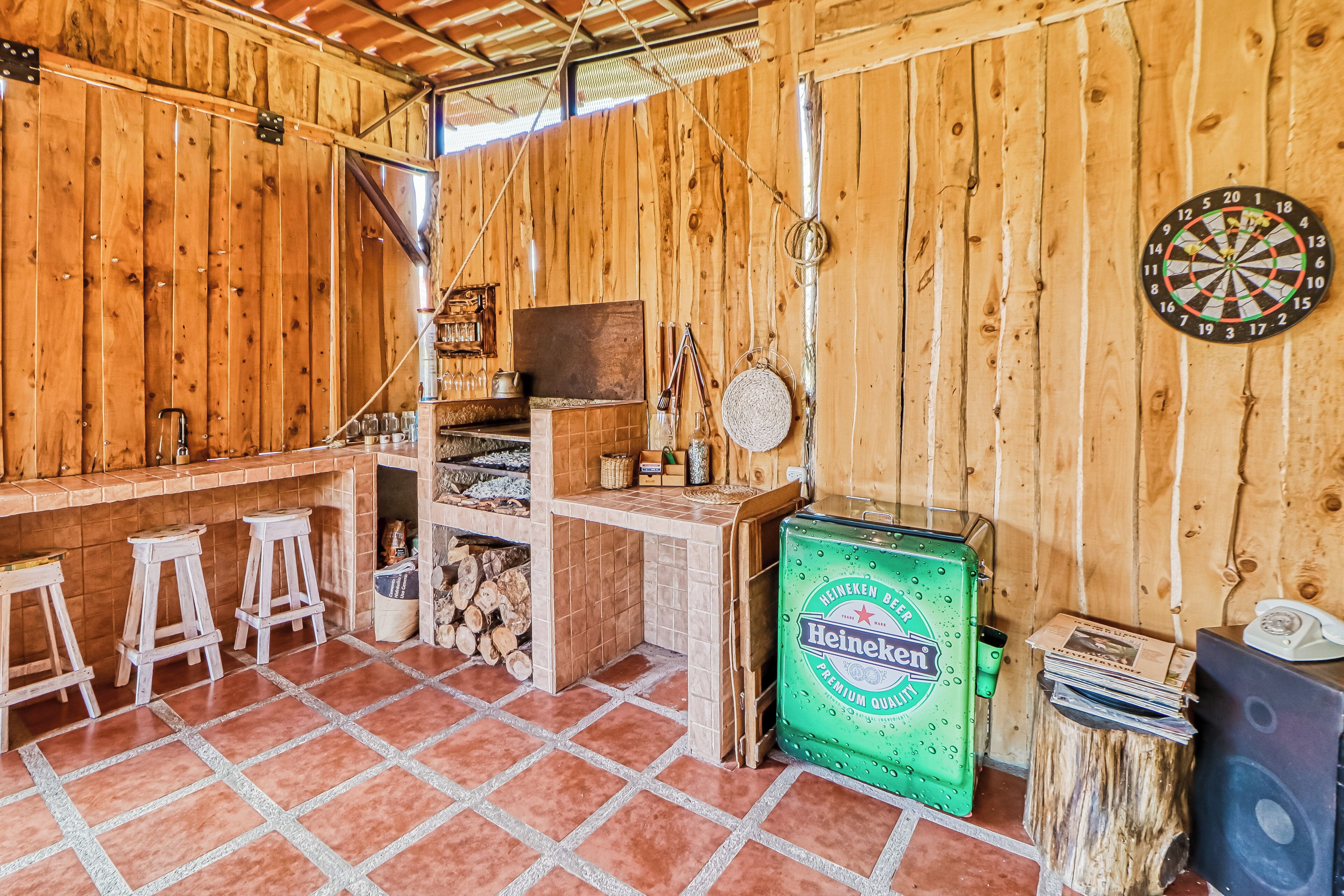 A rustic wooden room with a bar area, stools, a dartboard, and a Heineken cooler.