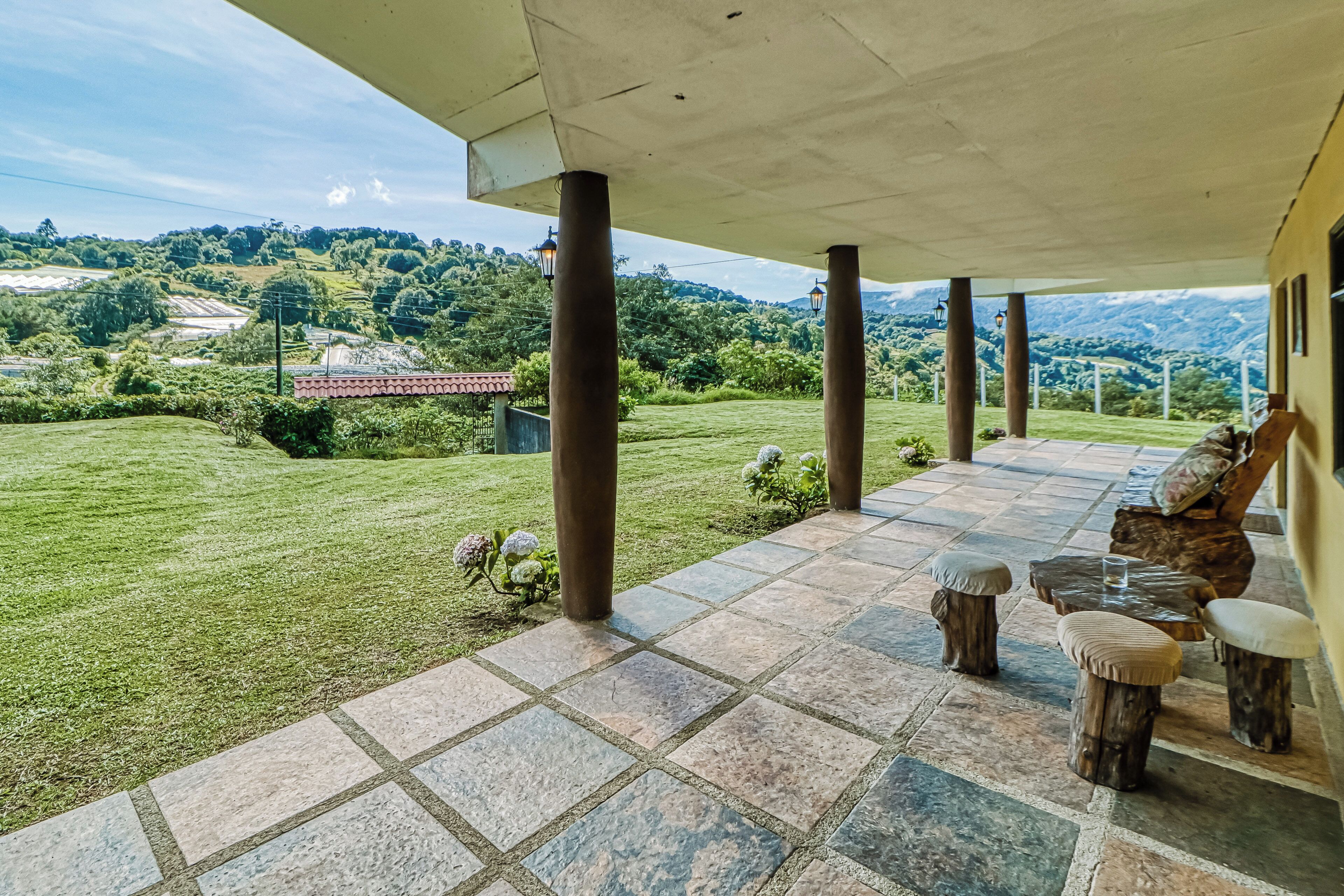 A covered outdoor area with stone flooring, wooden furniture, and a view of a green landscape with hills.