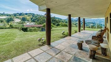 A covered outdoor area with stone flooring, wooden furniture, and a view of a green landscape with hills.