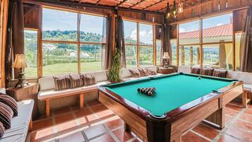 A pool table in a room with large windows, wooden beams, and a view of a green landscape.