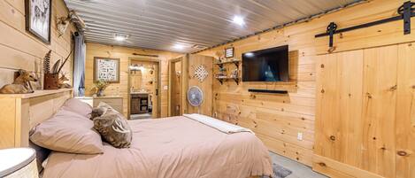 A wooden cabin interior with a bed, a television mounted on the wall, and various hunting-related decorations.