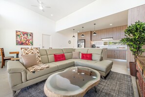 A modern living room with a sectional sofa, a coffee table, and a kitchen area with wooden cabinets and a refrigerator. - Playa Avellanas Villa: Modern Luxury, Pool, Views (Veintisiete de Abril)