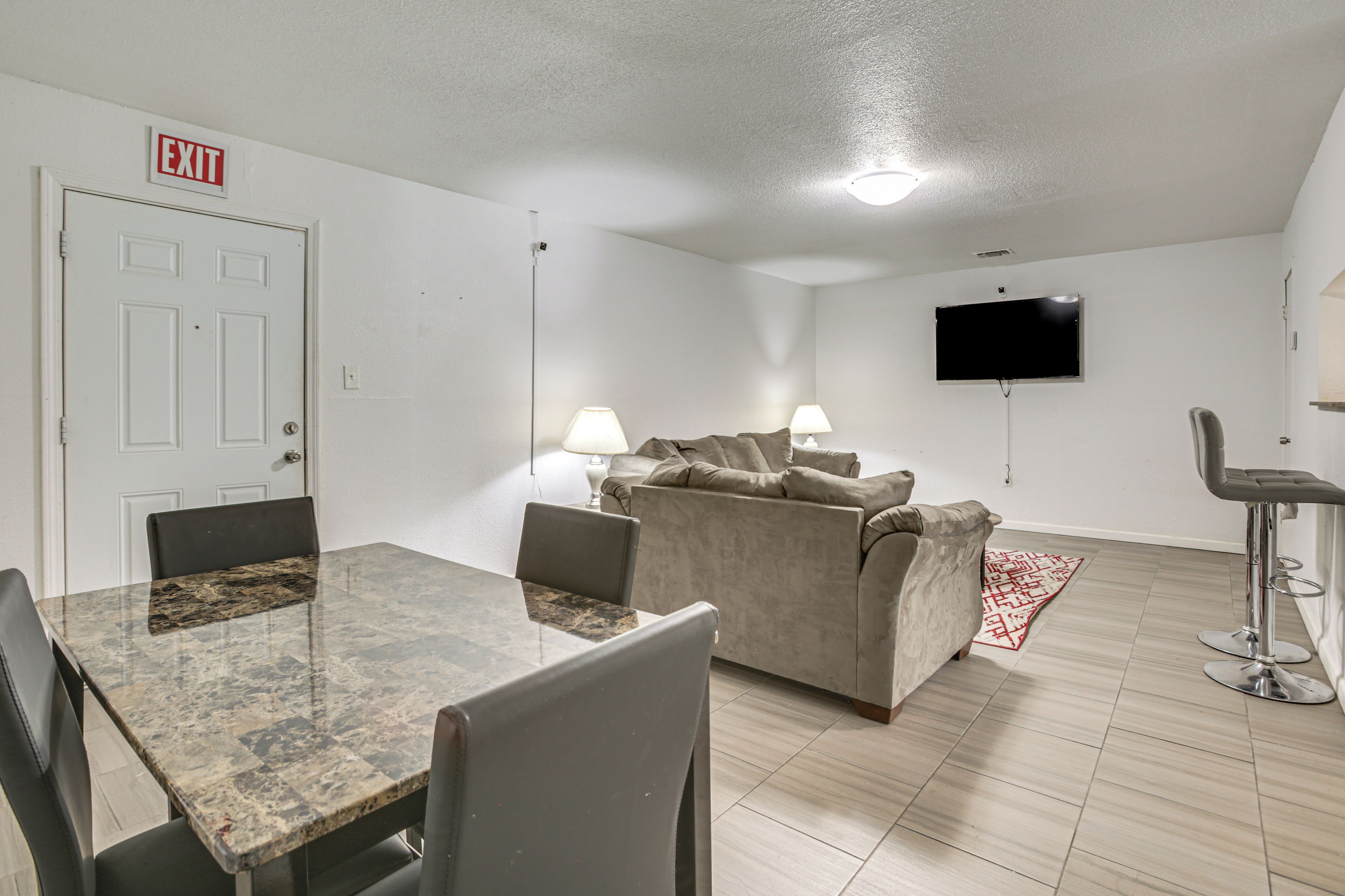 Una habitación con una mesa de comedor, sillas, un sofá, un televisor montado en la pared y un letrero de salida.