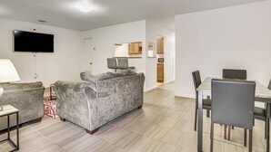 Uma sala de estar moderna com um sofá cinzento, uma TV de tela plana, uma mesa de jantar com cadeiras e uma área de cozinha ao fundo.