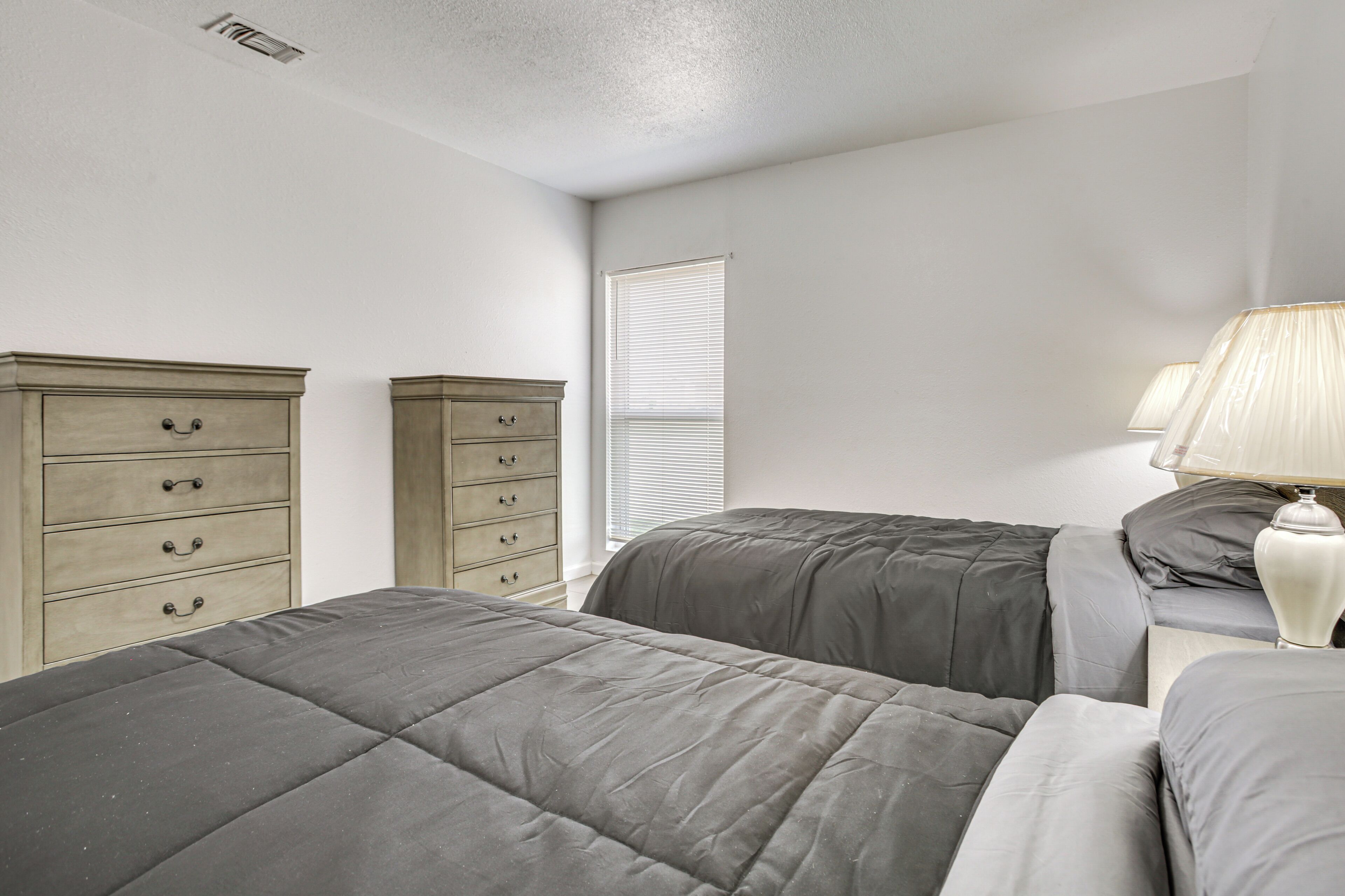 A bedroom with two beds, a dresser, a nightstand with a lamp, and a window with blinds.
