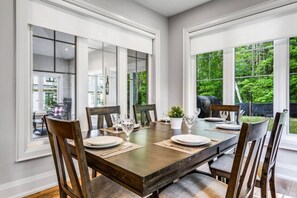 Comedor con una mesa grande dispuesta para seis comensales, rodeada de sillas y amplias ventanas que dan a vistas de vegetación.