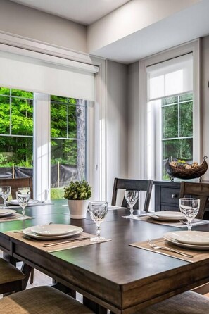 Een eetgedeelte met een houten tafel gedekt voor vier personen, grote ramen met uitzicht op groen en een decoratieve schaal op tafel.