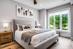 Une chambre dotée d’un grand lit, de deux tables de chevet, d’une fenêtre donnant sur un paysage verdoyant et d’une œuvre d’art représentant des plumes, accrochée au mur.