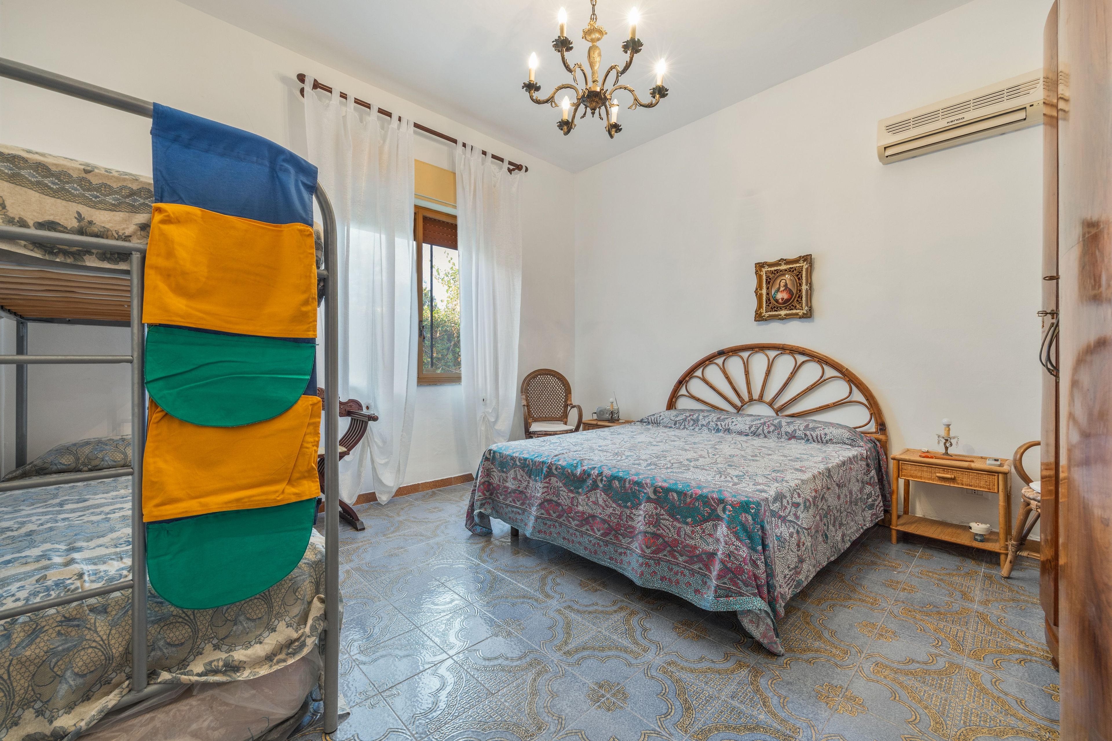 Una habitación con literas, una cama con colcha estampada, una silla de madera, una mesita de noche, aire acondicionado de pared y un candelabro.