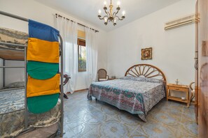 A room with bunk beds, a bed with a patterned bedspread, a wooden chair, a bedside table, a wall-mounted air conditioner, and a chandelier.