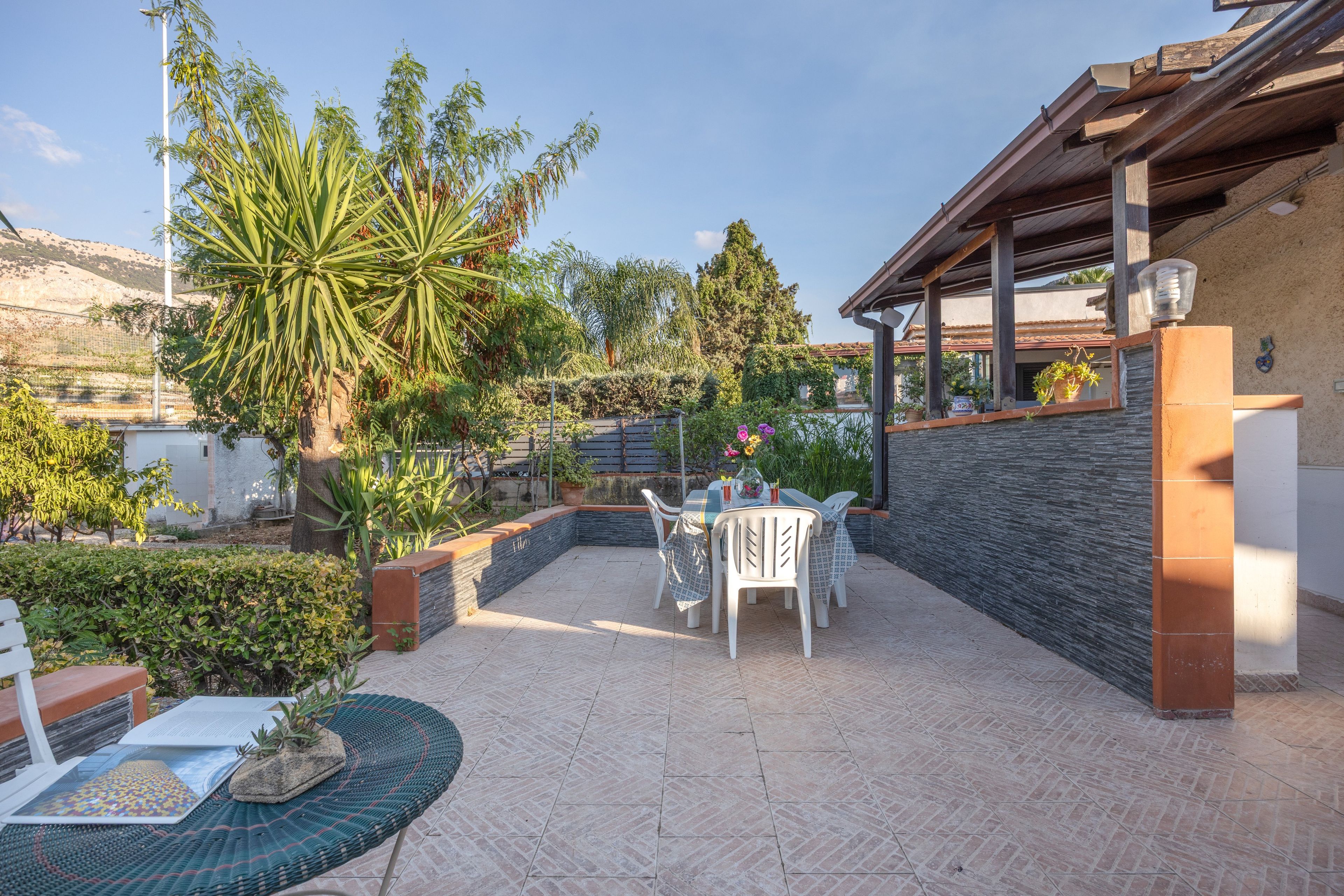 Eine Terrassenfläche mit Tisch und Stühlen, umgeben von Pflanzen und Blick auf einen Hügel.