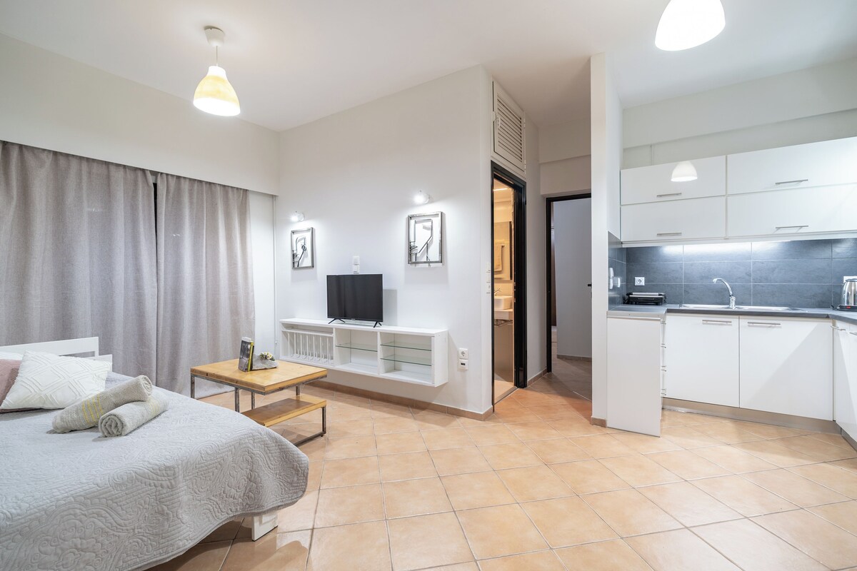 Ein modernes Hotelzimmer mit Bett, kleinem Tisch, Fernseher, Küchenzeile und einem durch eine offene Tür einsehbaren Badezimmer.
