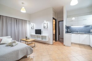Ein modernes Hotelzimmer mit Bett, kleinem Tisch, Fernseher, Küchenzeile und einem durch eine offene Tür einsehbaren Badezimmer.