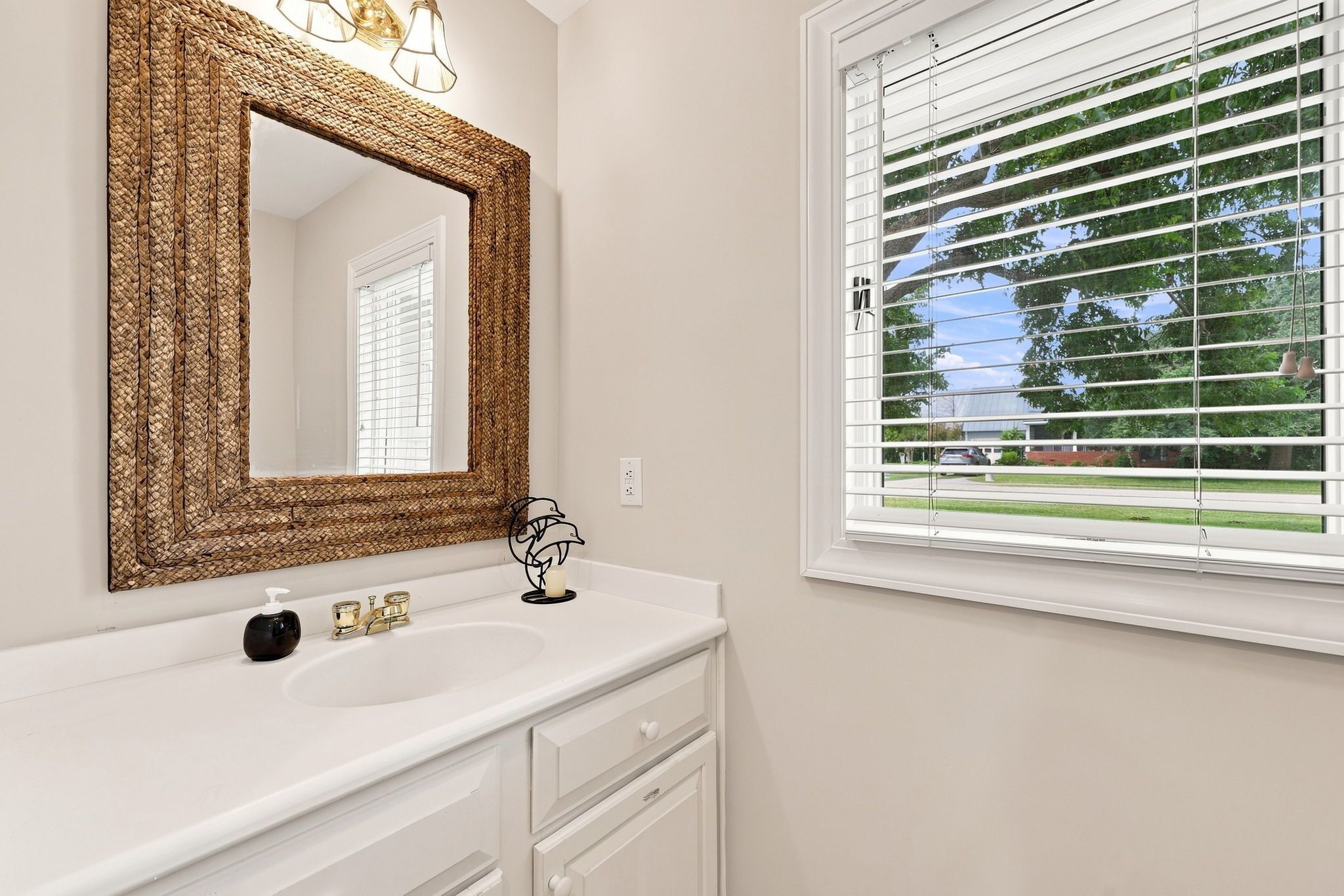 A bathroom with a white vanity, a large mirror with a woven frame, a sink with a soap dispenser, and a window with blinds.