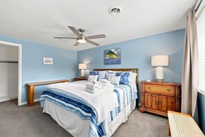 Ein Schlafzimmer mit einem Bett, einem Deckenventilator, einem Nachttisch, einer Lampe, einer Kommode und einem Fenster mit Vorhängen.