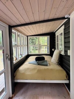 Une petite chambre fermée, avec un lit simple, de grandes fenêtres et un plafond en bois.