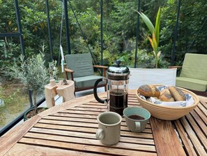 Une table en bois, une cafetière à piston, une tasse, un bol de pain et une chaise, le tout installé dans un jardin.