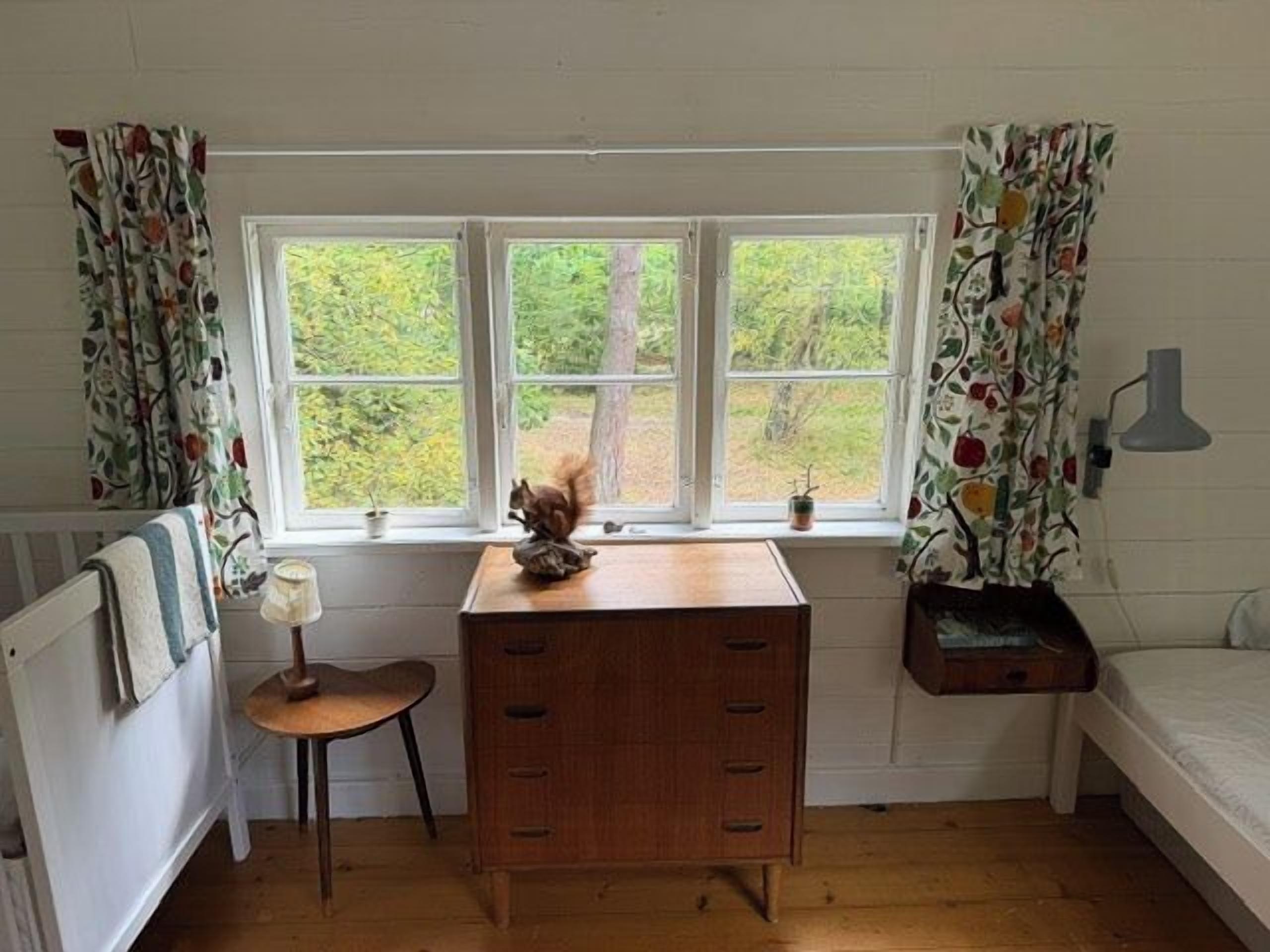A room with a wooden desk, a chair, a bed, a window with curtains, and a small table with a lamp.