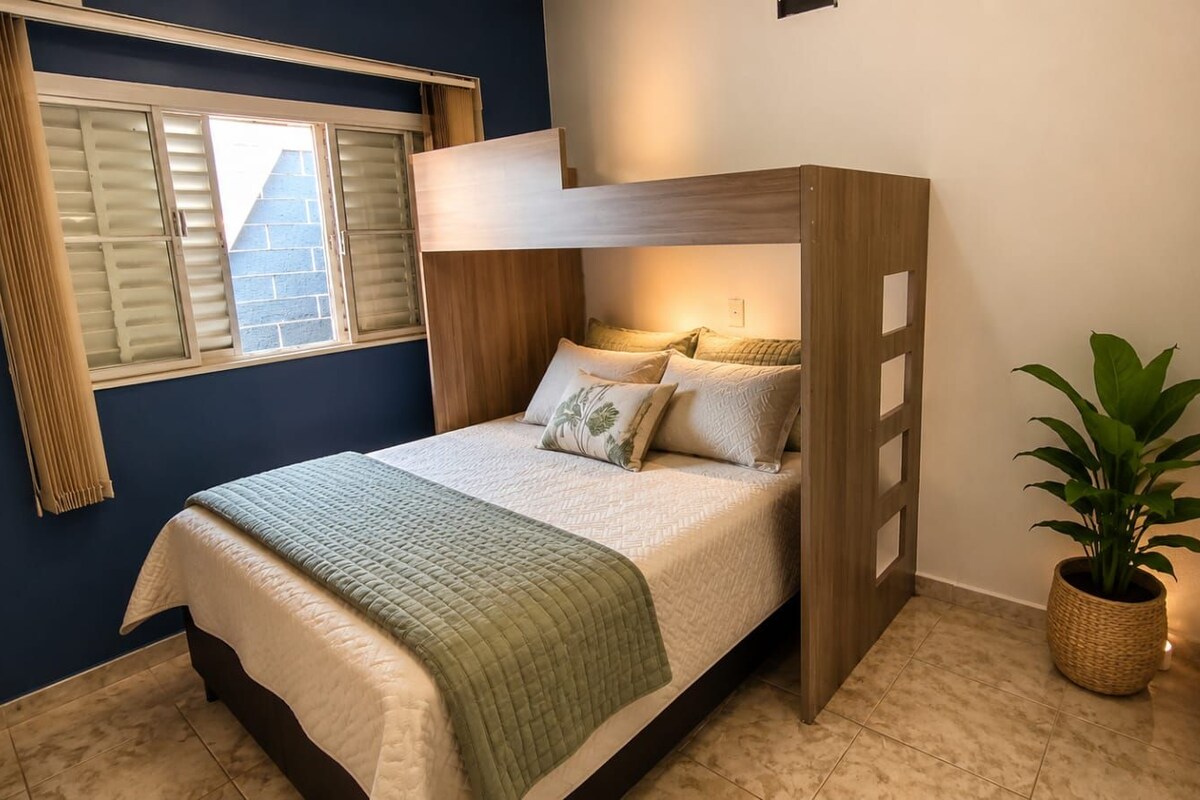 A bunk bed with a wooden headboard, a bedside table, a potted plant, and a window with blinds.