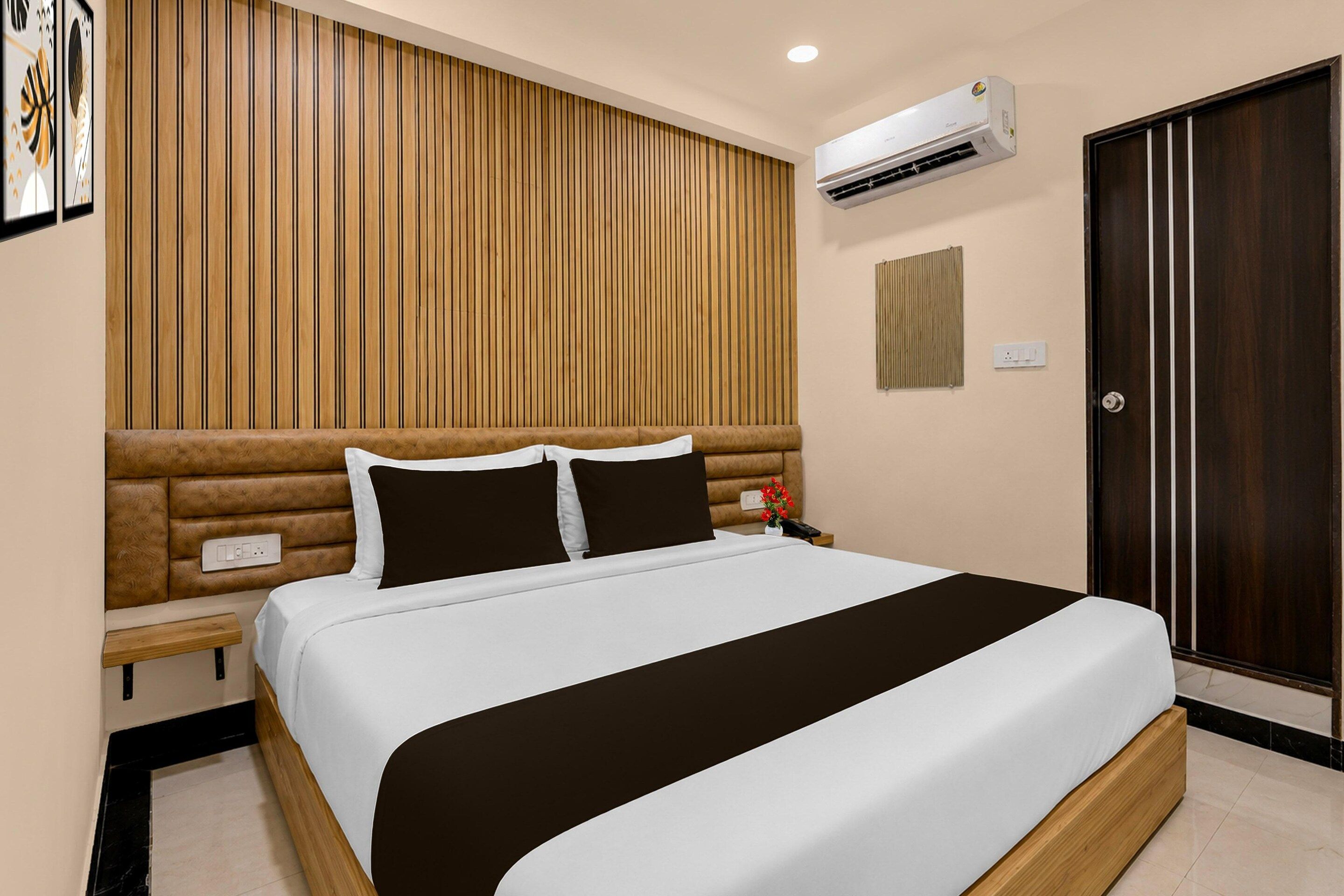 A hotel room with a large bed, a wooden headboard, an air conditioning unit, a bedside table, a nightstand, and a door.