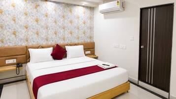 A hotel room with a bed, a bedside table, a red throw blanket, a wall air conditioner, and a dark wooden door.