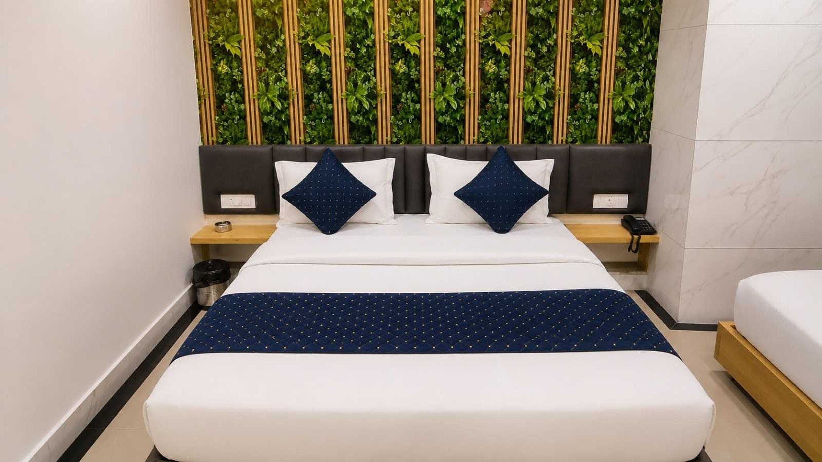 A hotel room with a bed, a bedside table with a phone, a trash bin, and a wall with vertical greenery.