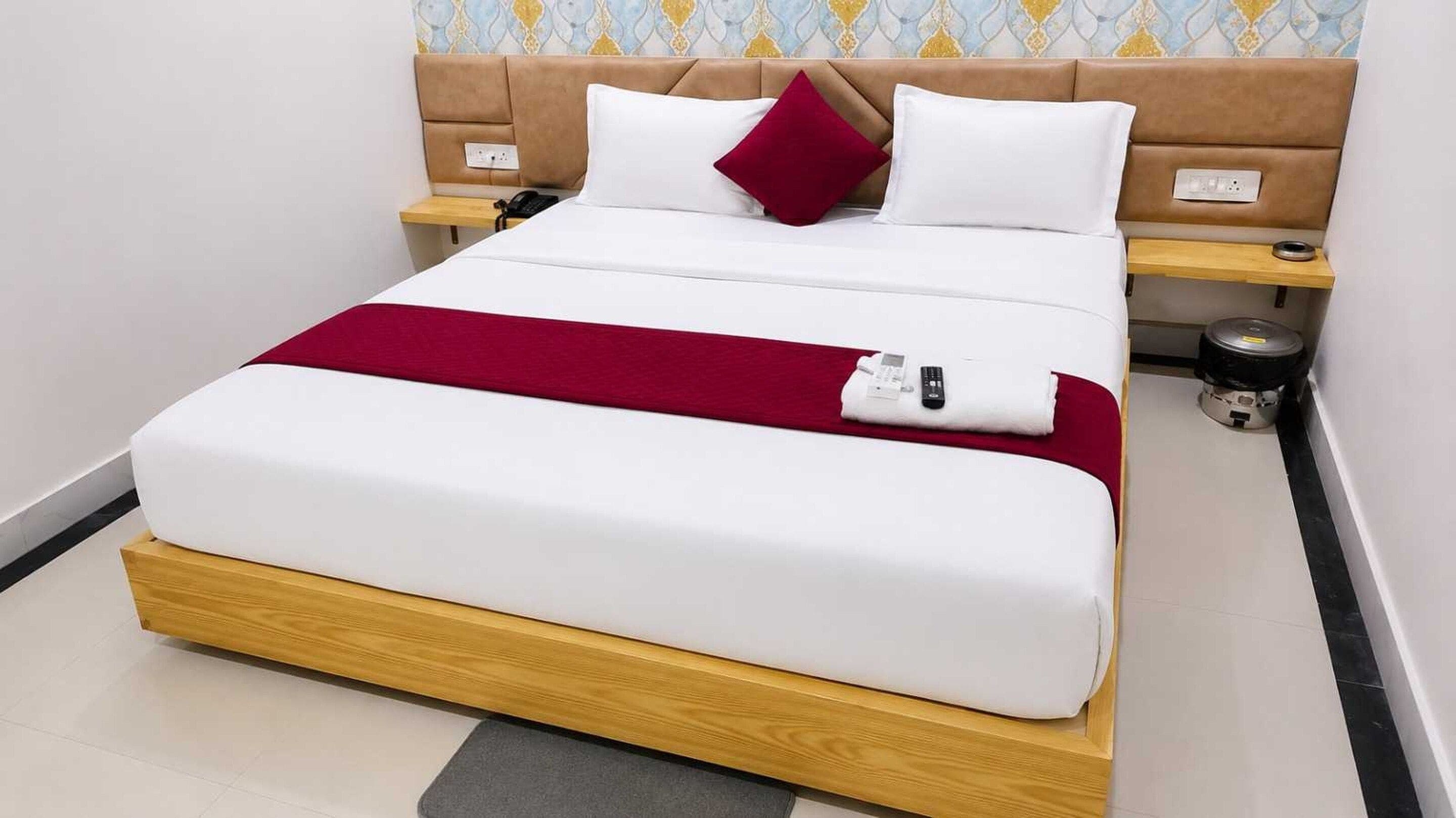 A hotel room with a bed, a bedside table, a nightstand, a red throw blanket, a white pillow, a brown headboard, and a wall with a patterned wallpaper.
