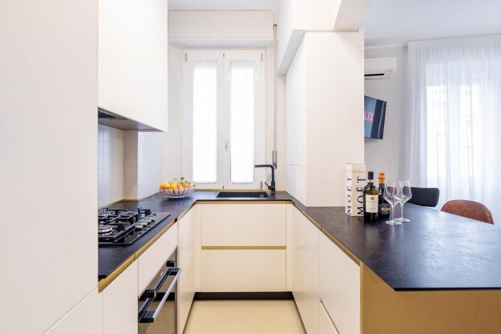 A modern kitchen with a black countertop, white cabinets, and a built-in oven. There is a window with white curtains and a flat-screen TV mounted on the wall.