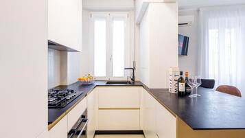 A modern kitchen with a black countertop, white cabinets, and a built-in oven. There is a window with white curtains and a flat-screen TV mounted on the wall.