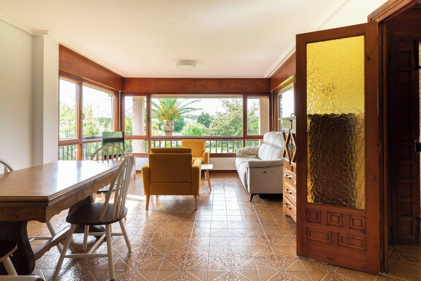 A spacious room with a wooden ceiling, a dining table with chairs, a sofa, and a view through large windows.