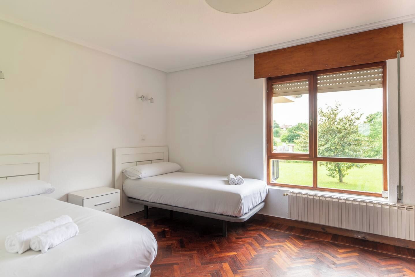 Une chambre d’hôtel avec deux lits, un plancher en bois, une fenêtre donnant sur un espace verdoyant et un radiateur.