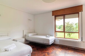 A hotel room with two beds, a wooden floor, a window with a view of greenery, and a radiator.