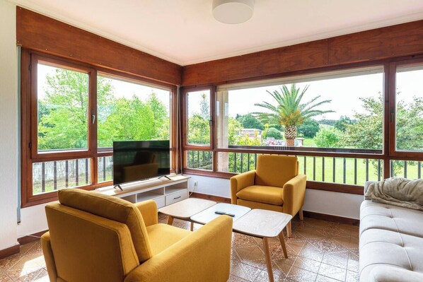 A living room with a TV, yellow armchairs, a white sofa, and large windows with a view of greenery.