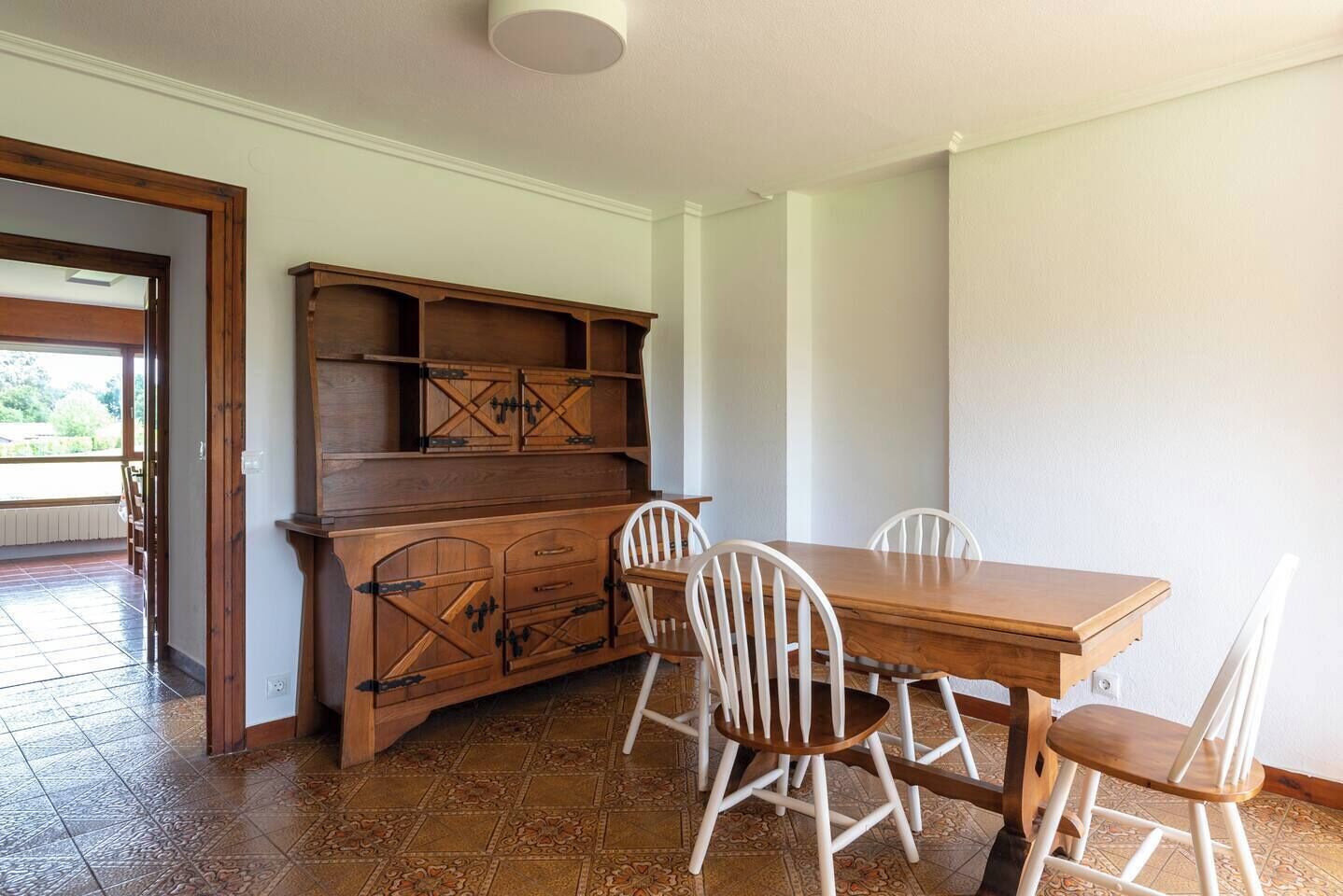 A dining area with a wooden table and white chairs, a wooden cabinet with drawers, and a tiled floor.