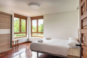 A bedroom with a large bed, wooden flooring, a radiator, and a window with a view of greenery.
