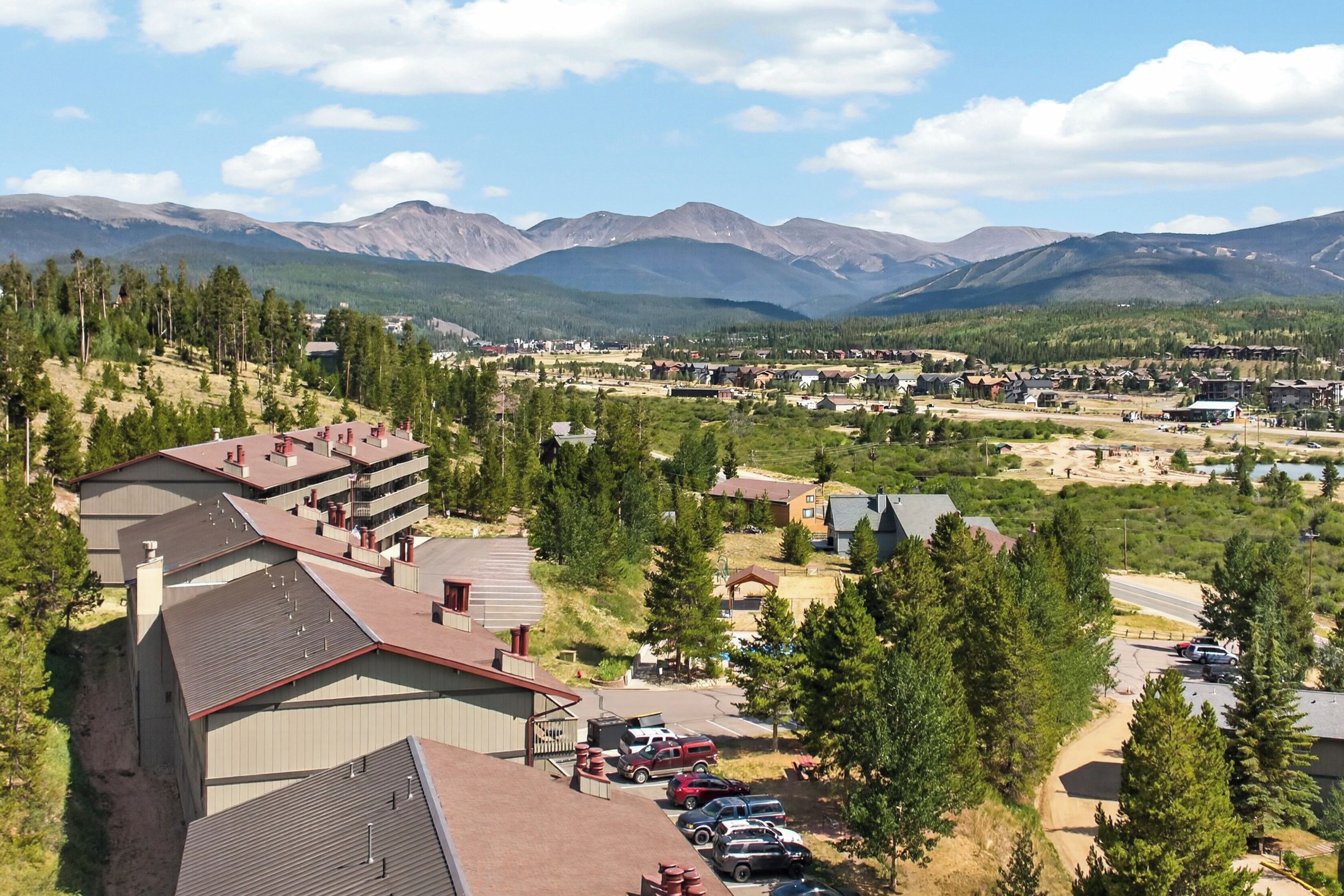 A residential area with a large building featuring multiple balconies, surrounded by dense forest and mountains in the background.