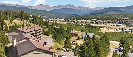 A residential area with a large building featuring multiple balconies, surrounded by dense forest and mountains in the background.