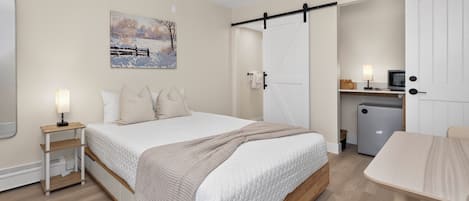 A bedroom with a bed, bedside tables, a painting, a wardrobe, a desk, and a refrigerator.