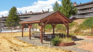 Un pabellón de madera con mesas de picnic, rodeado de grava y plantas, cerca de una piscina y edificios residenciales.