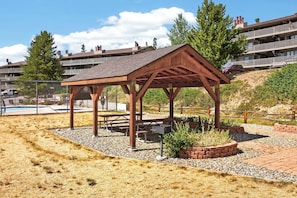 En træpavillon med picnicborde, omgivet af grus og beplantning, i nærheden af en swimmingpool og boligbygninger.