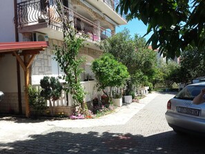 A residential street with a parked car, a building with a balcony, and a garden with potted plants. - Apartment Mediteran Okrug Gornji (Okrug Gornji)