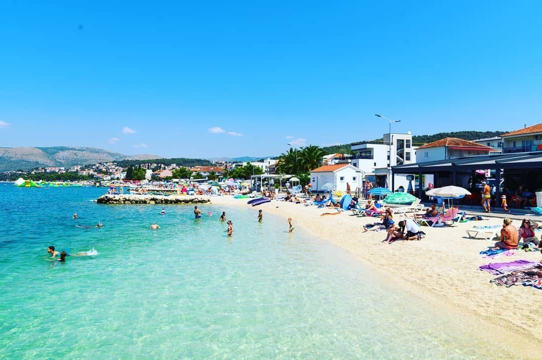 A beach with clear water, people swimming and sunbathing, and buildings in the background.