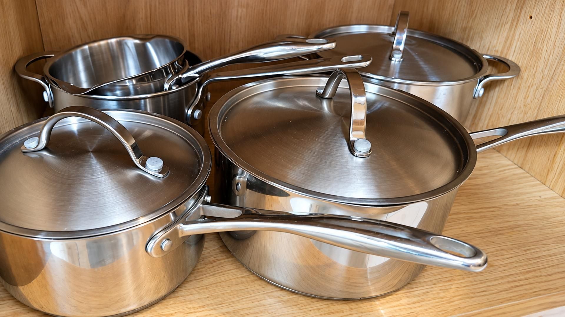 Edelstahlkochgeschirr mit Deckeln und Griffen, vermutlich zum Kochen, auf einem hölzernen Regal gelagert.