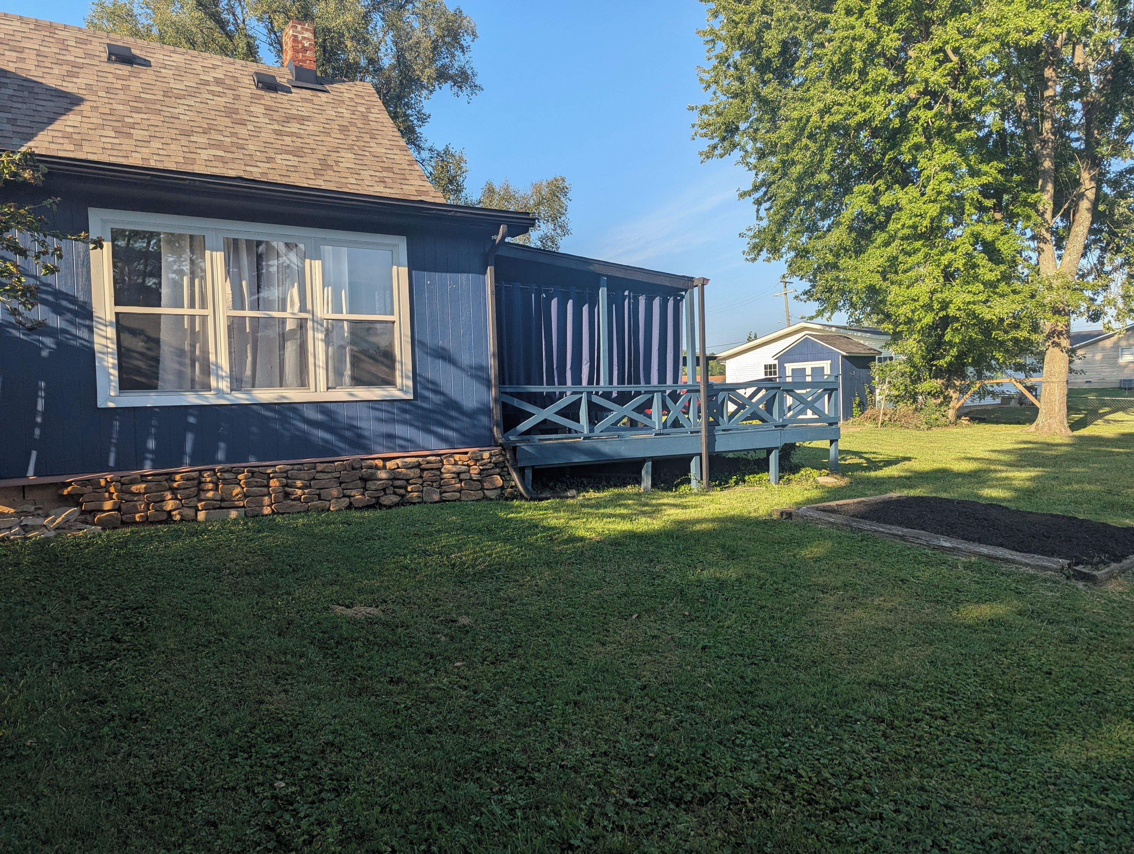 Een huis met een stenen fundering, een houten terras en een grote boom in de voortuin.