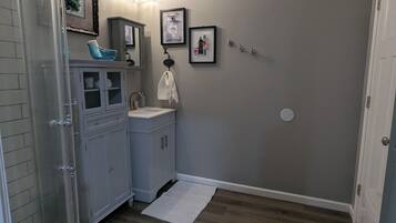 Een badkamer met douche, toilet, wastafel en opbergmeubel. Er liggen twee vloerkleden op de vloer, een handdoek hangt aan de muur en er zijn afbeeldingen in lijsten.