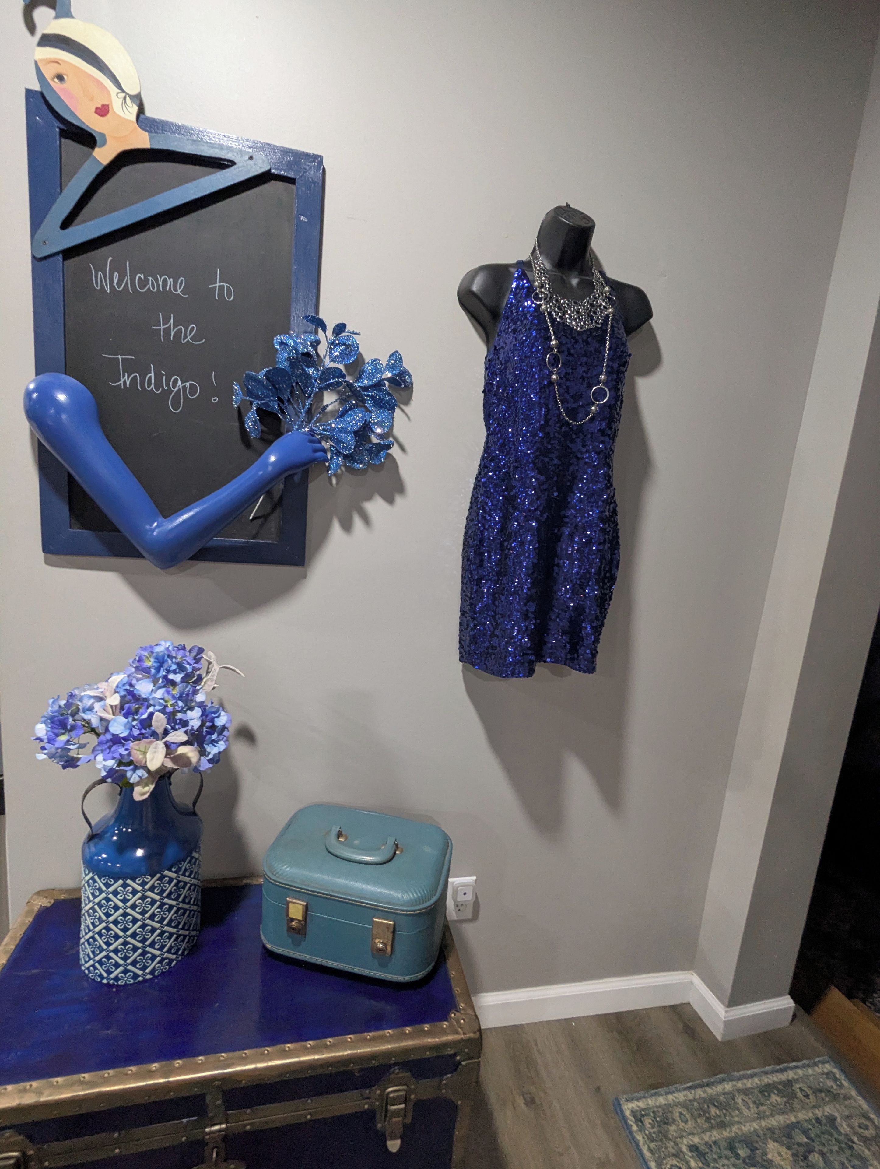 Una habitación con una pizarra, un maniquí vestido de azul, un florero con flores y una maleta.