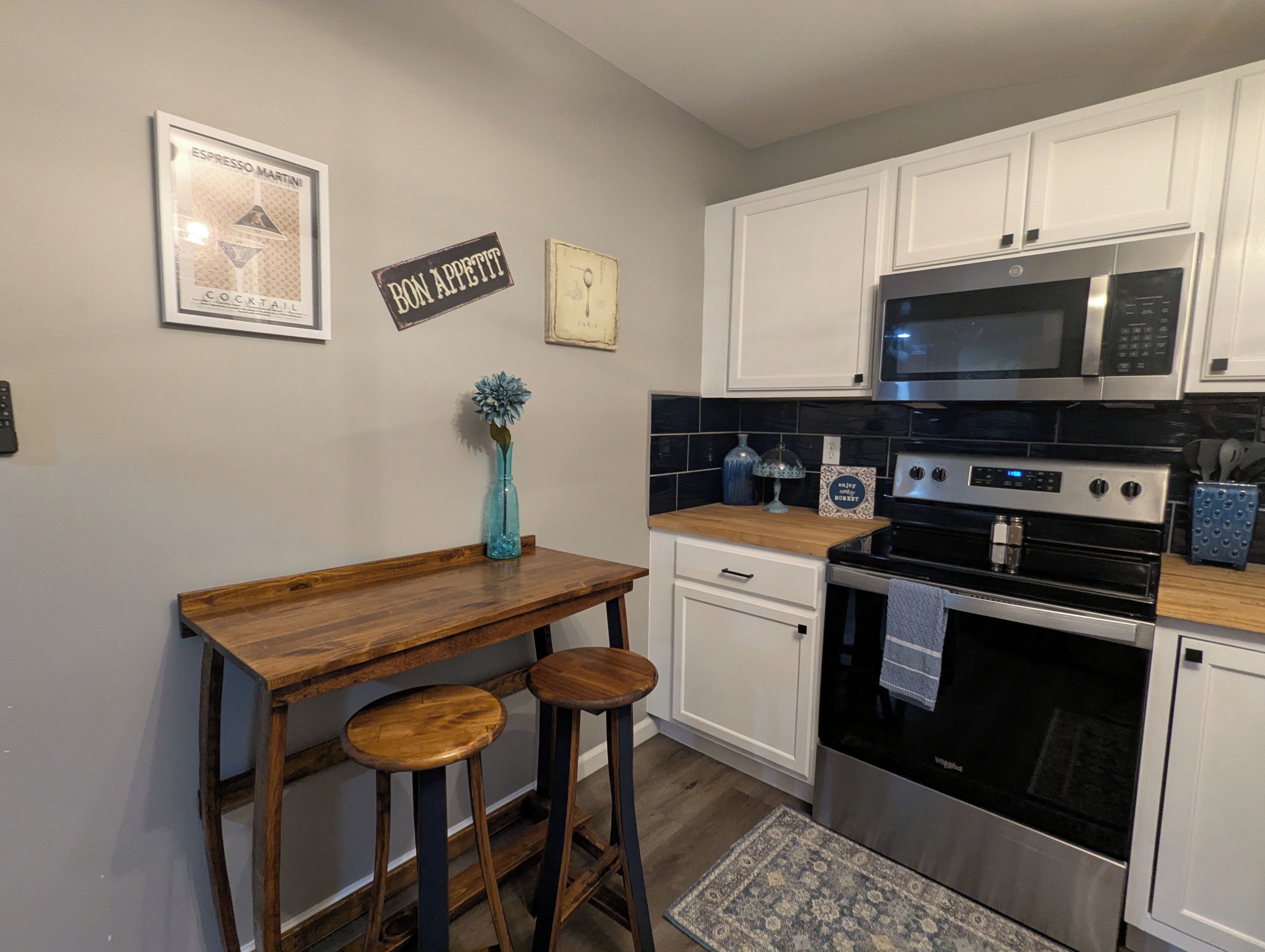 Una cocina con armarios blancos, una barra de madera, dos taburetes, un jarrón azul y un microondas.