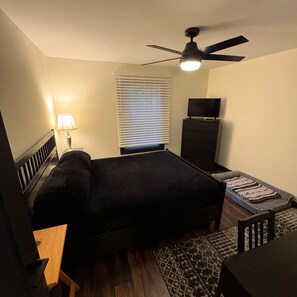 Ein Schlafzimmer mit Bett, Schreibtisch, Stuhl, Fernseher, Deckenventilator und einem Fenster mit Jalousien.