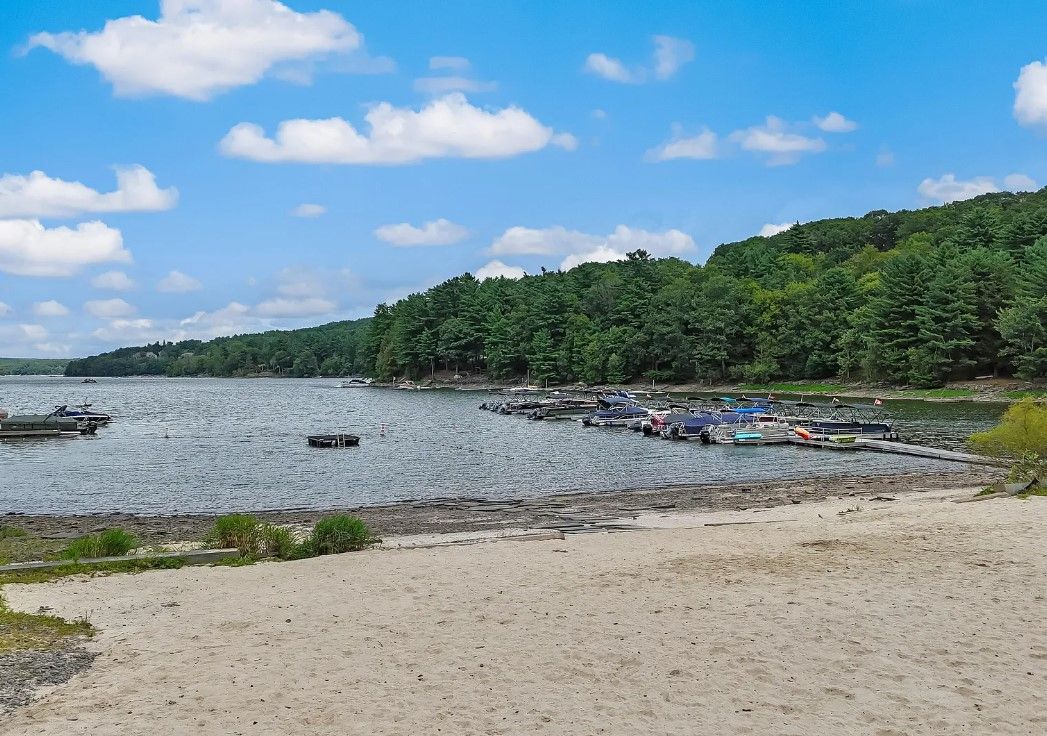 Blick auf einen See mit vertäuten Booten, einen Sandstrand und einen bewaldeten Hügel im Hintergrund.